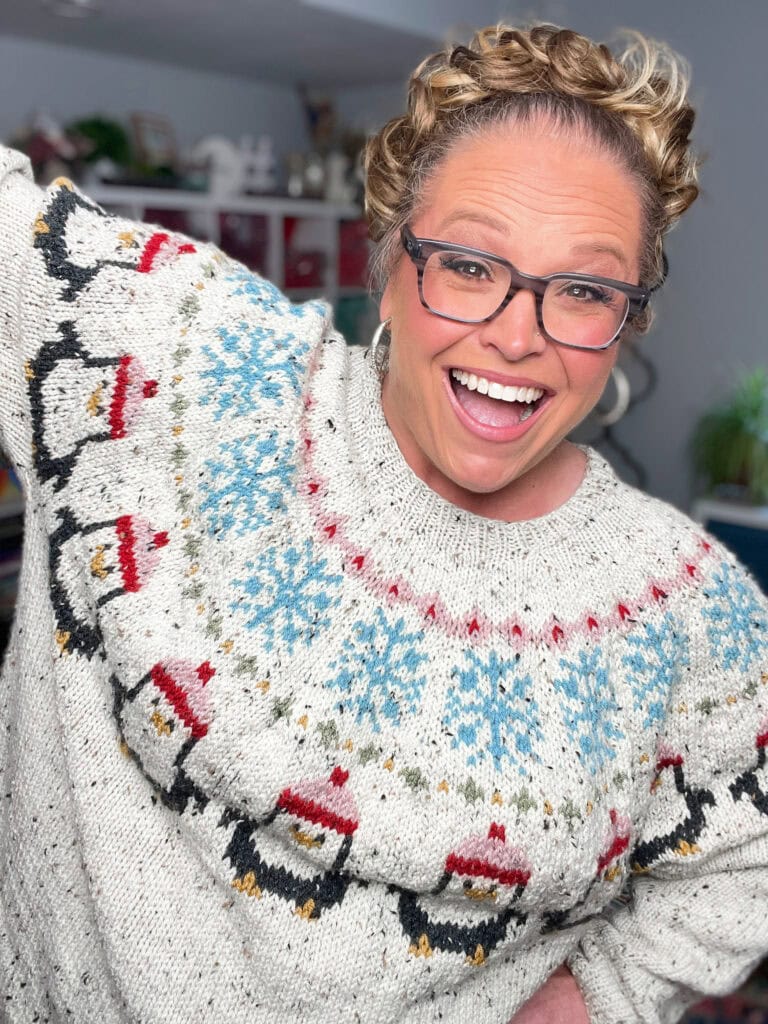 A smiling person with curly blonde hair and glasses wearing a white stranded colorwork sweater featuring penguins in red hats and blue snowflakes, standing indoors with shelves and plants in the background. -Marly Bird