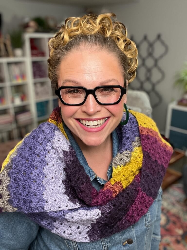A smiling person with curly blonde hair, wearing black glasses, a colorful crocheted shawl, and a denim shirt, poses indoors with shelves and a plant in the background. -Marly Bird