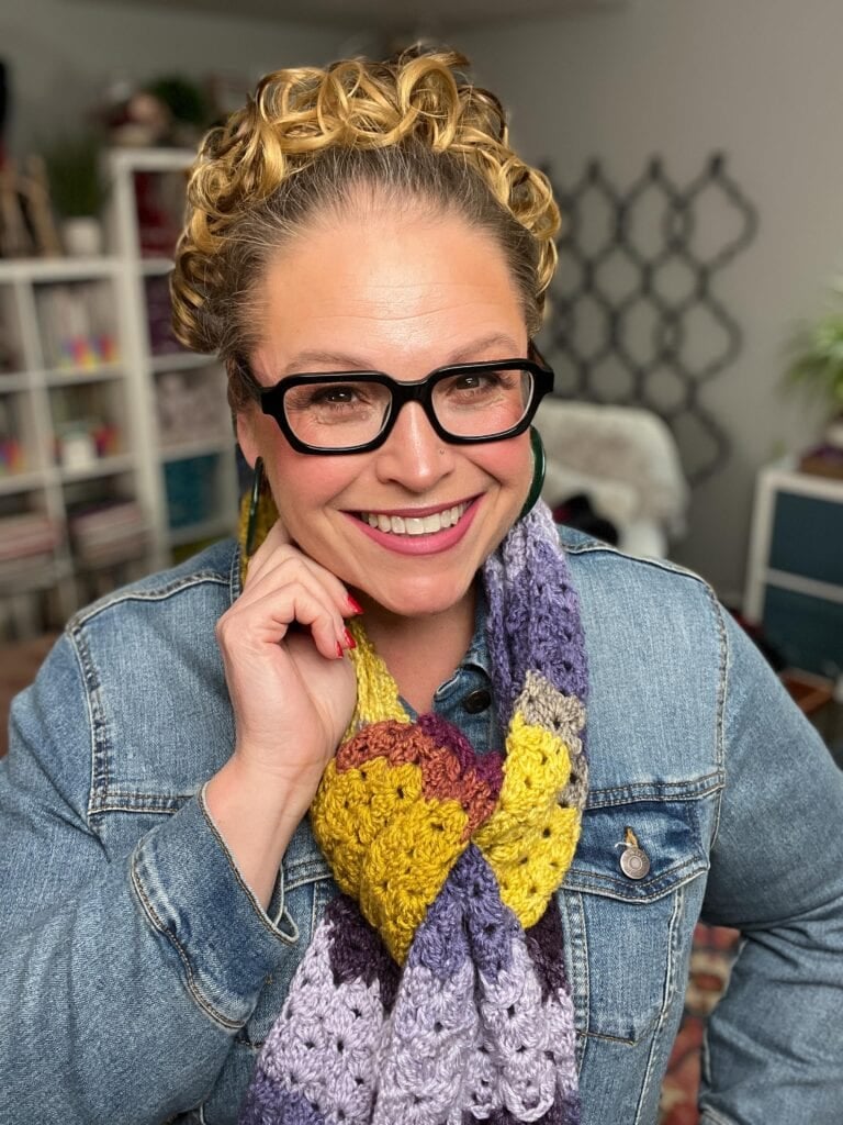 A smiling person with curly hair, glasses, and hoop earrings wears a denim jacket and a colorful crocheted scarf in shades of purple, yellow, and maroon, standing indoors with shelves and decor in the background. -Marly Bird