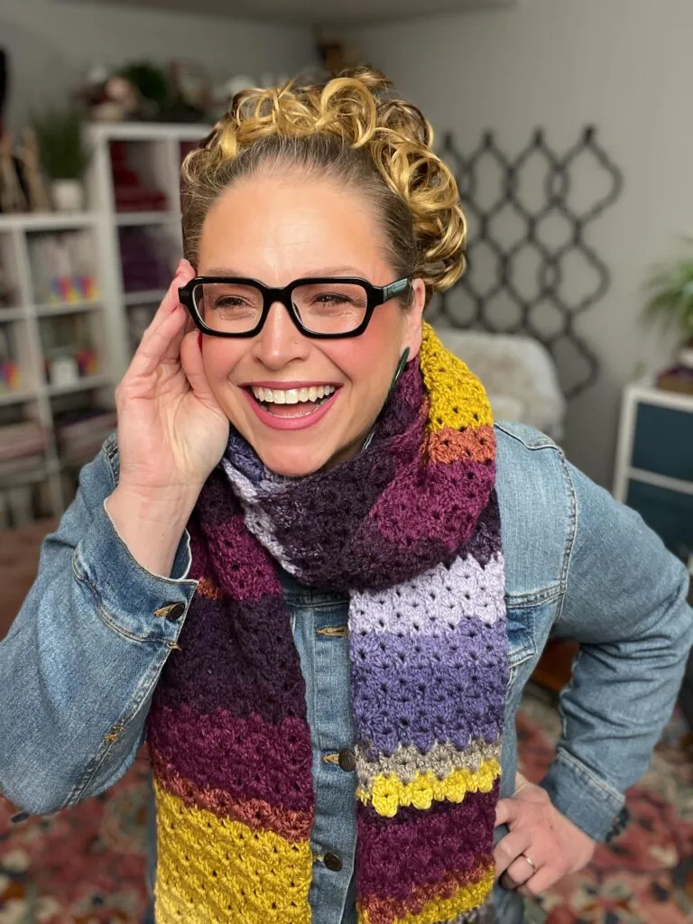 A smiling person with curly blonde hair and glasses wears a multicolored crocheted scarf and a denim jacket, standing indoors in a cozy, well-decorated room with shelves and plants in the background. -Marly Bird