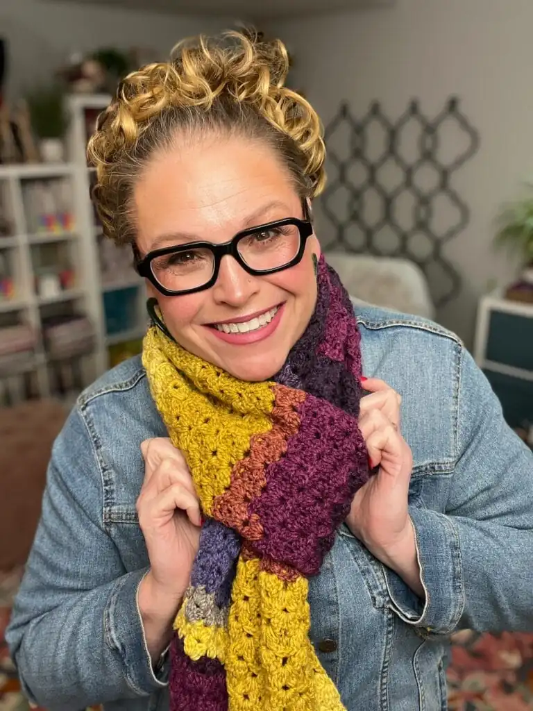 A smiling person with curly blonde hair, black glasses, and a denim jacket holds a colorful crocheted scarf in shades of yellow, purple, and maroon in a cozy, well-lit room. -Marly Bird