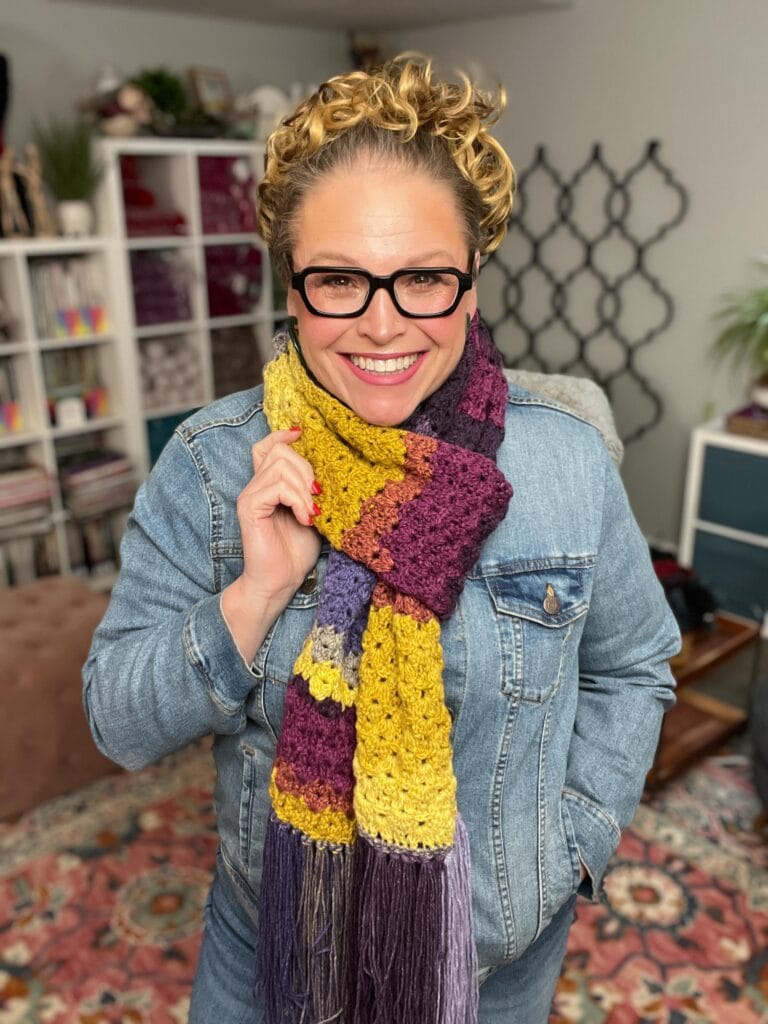 A smiling person wearing glasses, a denim jacket, and a colorful crocheted scarf stands indoors, with bookshelves and a decorative rug in the background. -Marly Bird