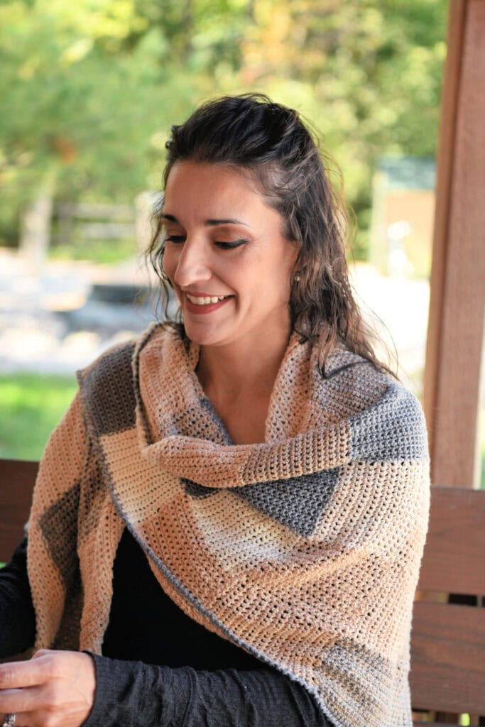 Beige and gray single crochet entrelac shawl draped over shoulders, showing textured blocks; worn outdoors on a wooden bench.