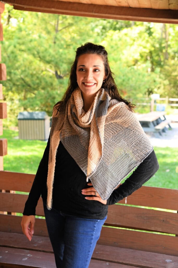 A knit shawl in beige and gray blocks drapes over a black top, showing clean stitch lines. Worn outdoors on a sunny porch.