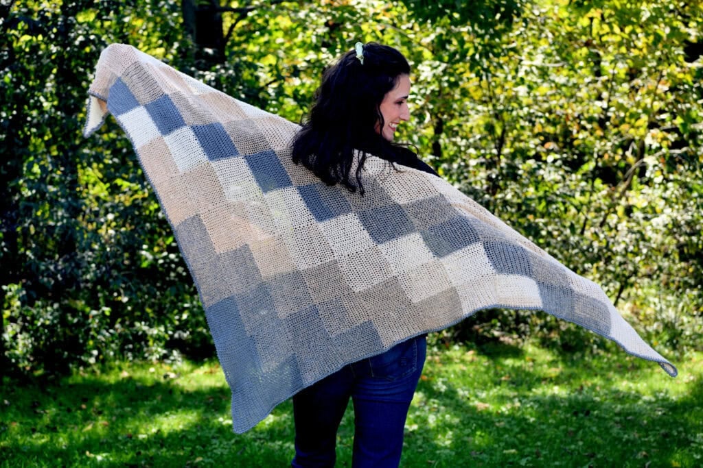 A woman models a large patchwork knit shawl with beige, gray, and cream squares on grass with leafy trees in the background.
