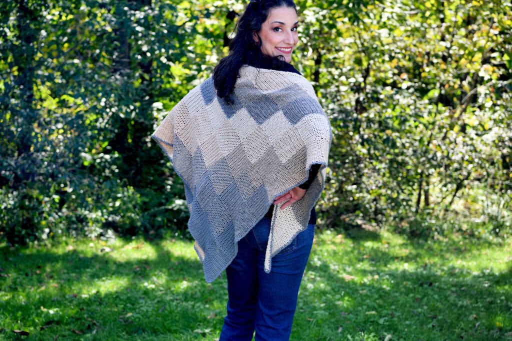 A crochet entrelac wrap in neutral tones is worn over blue jeans, highlighting its textured stitches outdoors in bright sunlight.