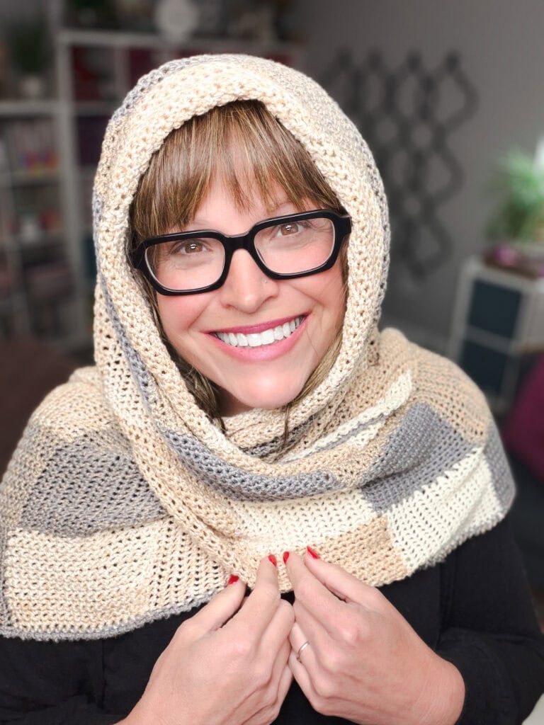 A woman models a striped, knit entrelac wrap scarf, showing its textured crochet pattern and soft drape over her head and shoulders.