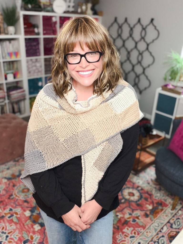 Woman models the beige and gray knit Pieces of You Wrap shawl, showing off its soft texture in a cozy, colorful room.