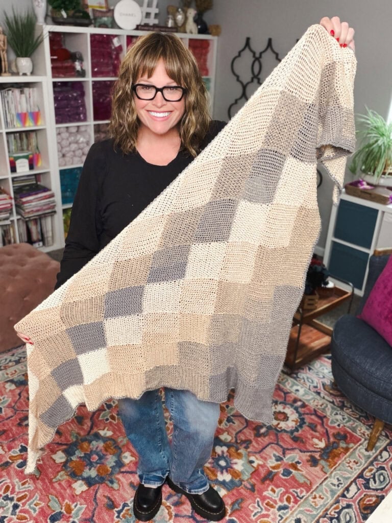 A smiling woman with glasses holds up a large, hand-knit blanket made of beige, tan, and gray squares in a cozy, colorful room.