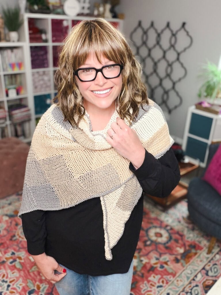 A woman models a neutral-tone crochet entrelac wrap, showing textured blocks, worn over black in a colorful room.