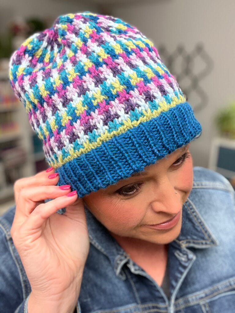 A person wearing a colorful knit hat with a blue brim and mosaic slip stitch pattern touches the side of the hat. The person has pink nail polish and is dressed in a denim jacket. Shelves are visible in the blurred background. -Marly Bird