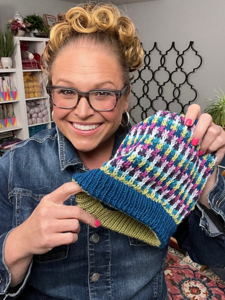 A smiling person with glasses and curly hair holds up a colorful knit hat, featuring a green brim and Mosaic Slip Stitch, wearing a denim jacket and showing off bright pink nail polish in a cozy, yarn-filled room. -Marly Bird
