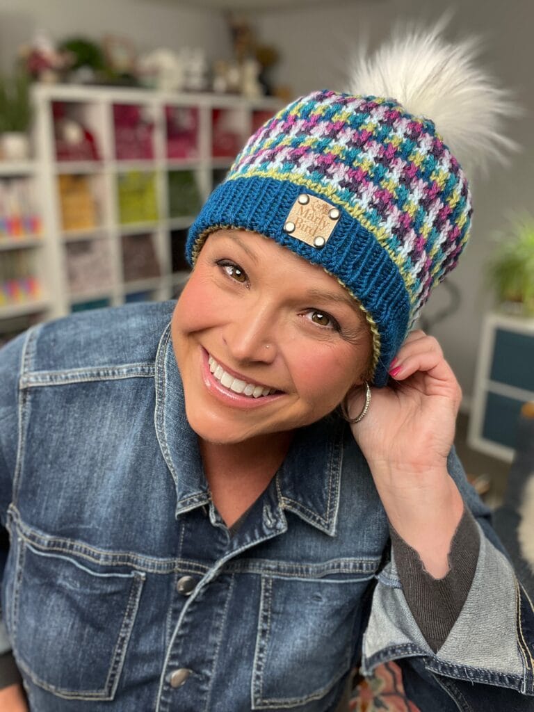 Smiling woman in a denim jacket holds the brim of a colorful, knitted beanie with a fluffy white pom-pom—perfect for stash-busting—standing in a cozy room with shelves of yarn in the background. -Marly Bird