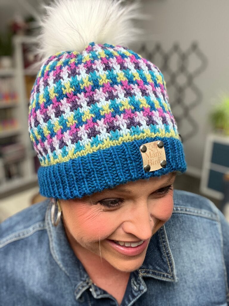 A smiling person wears a colorful stash-busting knit hat with a fluffy white pom-pom, blue brim, and "Wooly BIRD" label. They're dressed in a denim jacket. -Marly Bird