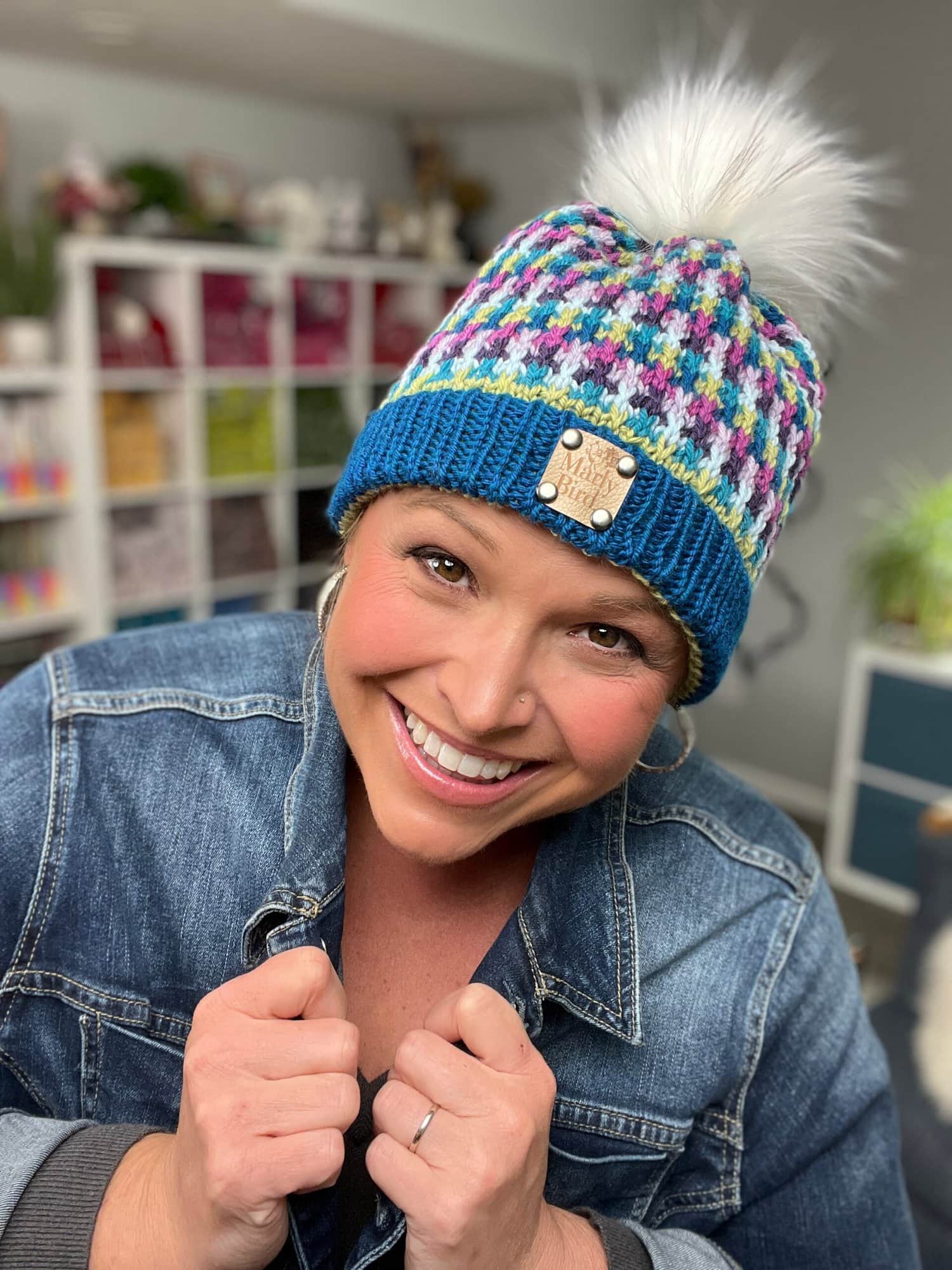 A smiling person in a denim jacket and colorful knit beanie with a white pom-pom poses indoors, holding their jacket collar. Shelves and plants are visible in the background—a cozy scene perfect for showcasing stash-busting knit hat patterns. -Marly Bird