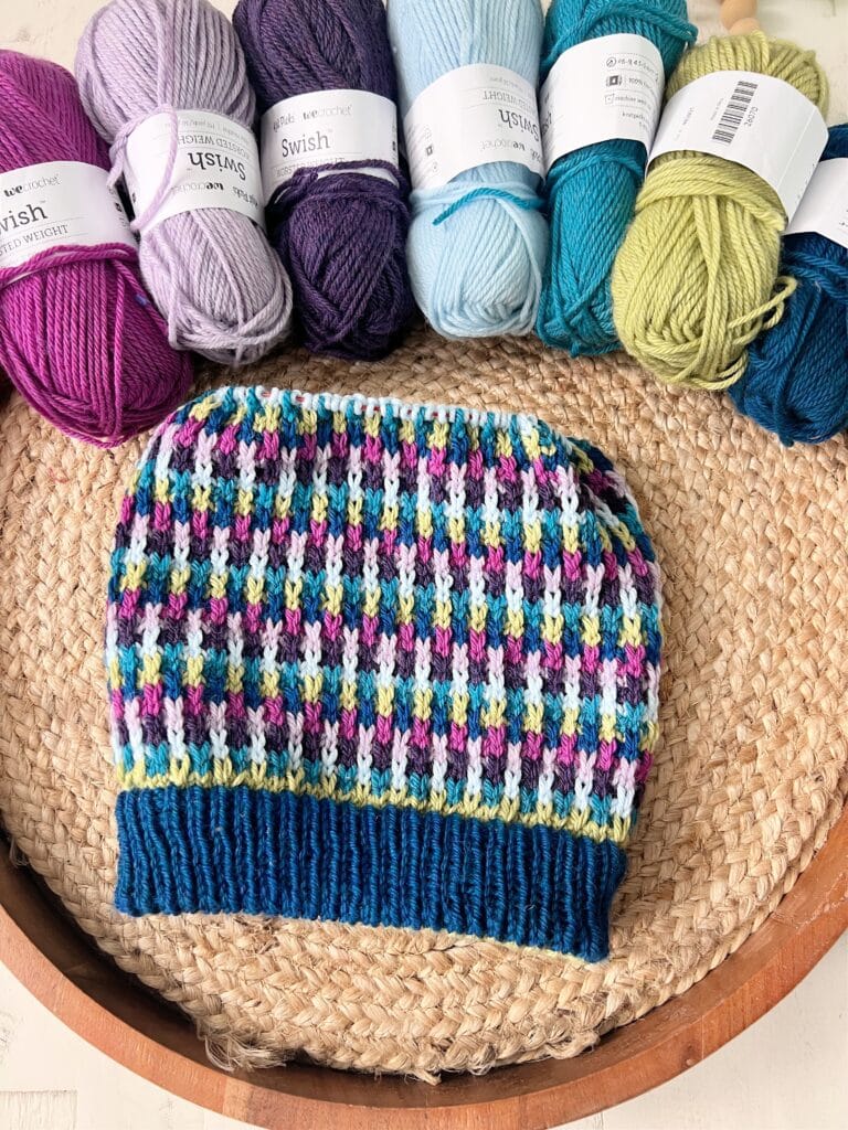 A colorful knit hat featuring a mosaic slip stitch geometric pattern is displayed on a woven mat, surrounded by skeins of yarn in purple, teal, green, blue, and white. -Marly Bird