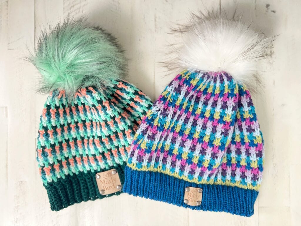 Two colorful knit beanies with large faux fur pom-poms on top. Inspired by the Flicker and Flash crochet hat, each features ribbed brims, leather tags, and vibrant hues—perfect for those seeking a fun accessory or a free crochet pattern idea. -Marly Bird