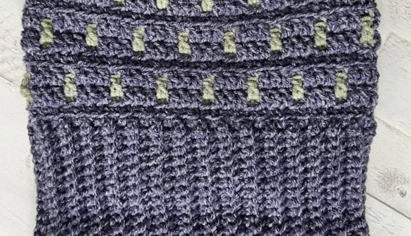 Close-up of a crocheted fabric showcasing a textured pattern. The bottom section consists of dark purple knit with diagonal ribbing, while the top has lighter green rectangular accents in horizontal rows. Perfect for a cozy Game Day look against a light, textured background. -Marly Bird