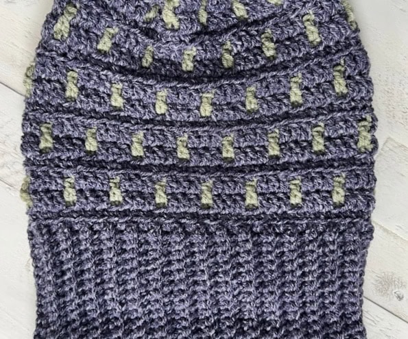 A crocheted beanie with a textured pattern featuring horizontal ridges and a checkered design, perfect for game day. It comes in a striking purple with light green accents and is stylishly displayed on a light wooden surface. -Marly Bird