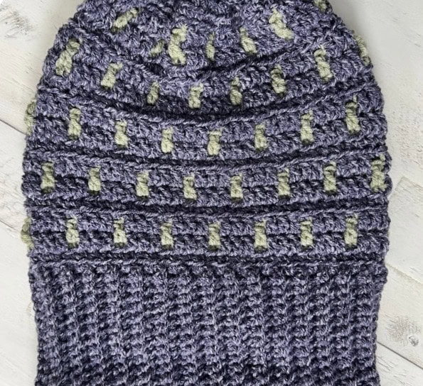 Hand-crocheted purple beanie with a textured, lattice-like pattern featuring small green accents, perfect for Game Day. This stylish crochet piece is displayed on a light grey wooden surface. -Marly Bird