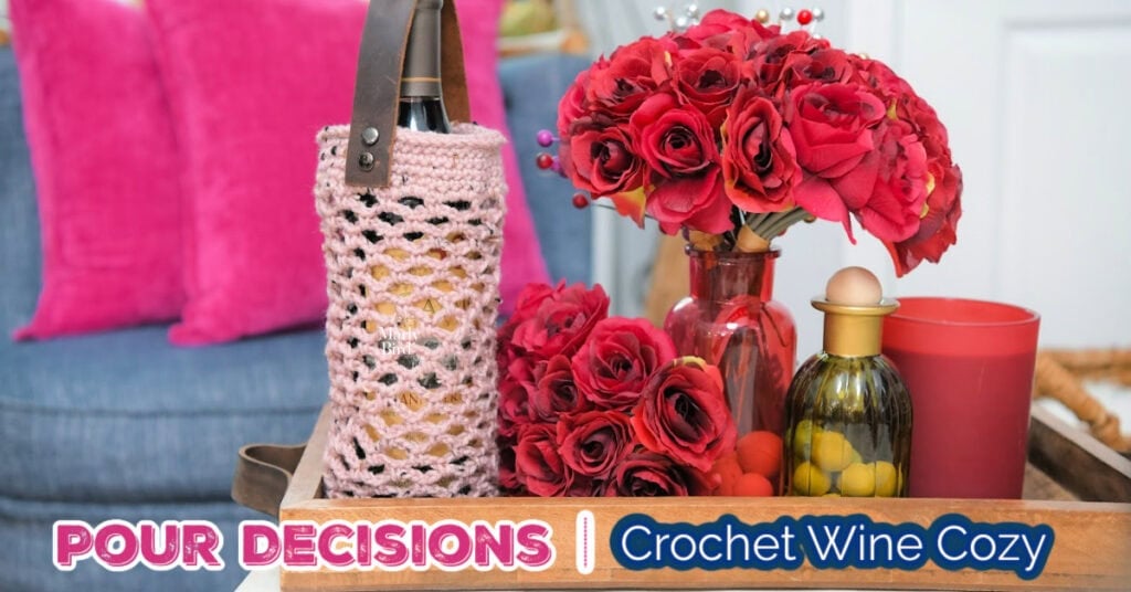Pink crochet wine cozy with leather strap on a tray with flowers, candle, and glass bottle; text: “POUR DECISIONS.”.