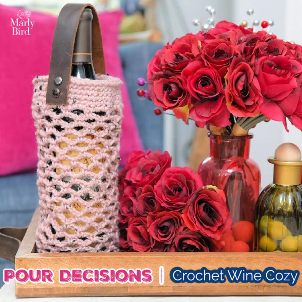 Pink crochet wine cozy with a leather strap wraps a wine bottle, shown on a tray beside red roses and a jar of olives.