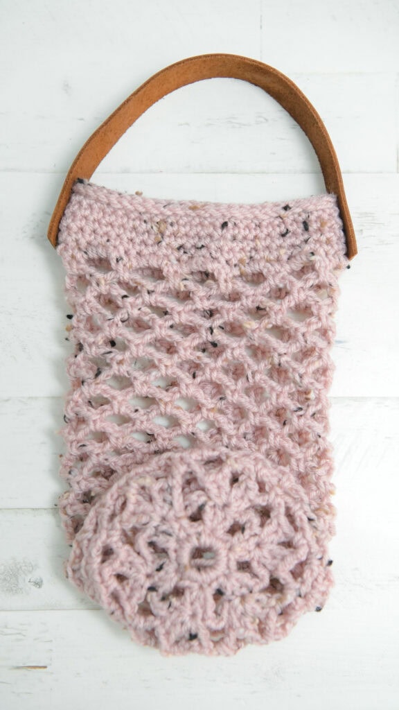 Light pink crocheted tote bag with open-weave stitch, brown leather strap, and attached circular pouch on white wood surface.