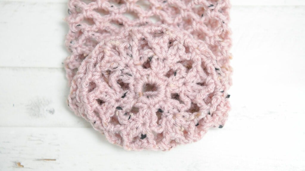 Light pink lacy crochet fabric with circular floral motifs, shown close-up on a white wood surface, highlighting stitch detail.