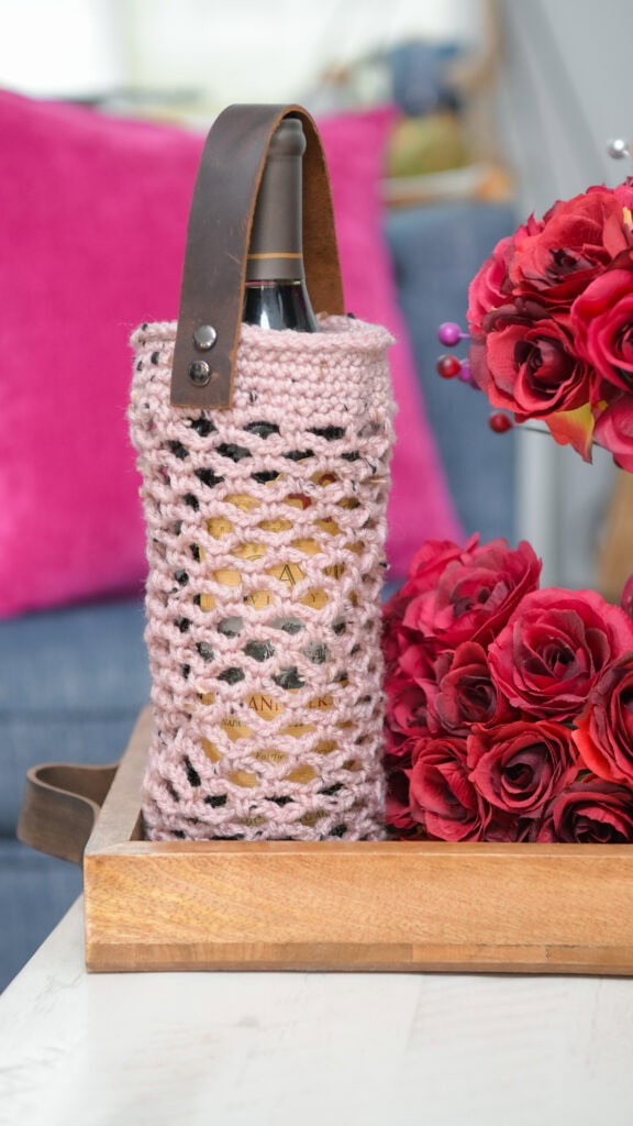 Pink crochet wine cozy with leather strap shown on bottle; textured stitches and neat finish, displayed on a wooden tray.