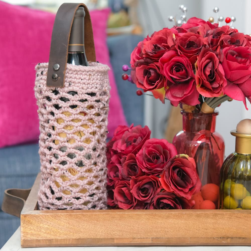 A pink crochet wine cozy with a leather strap covers a wine bottle on a tray, showcasing textured stitches and cozy detail.