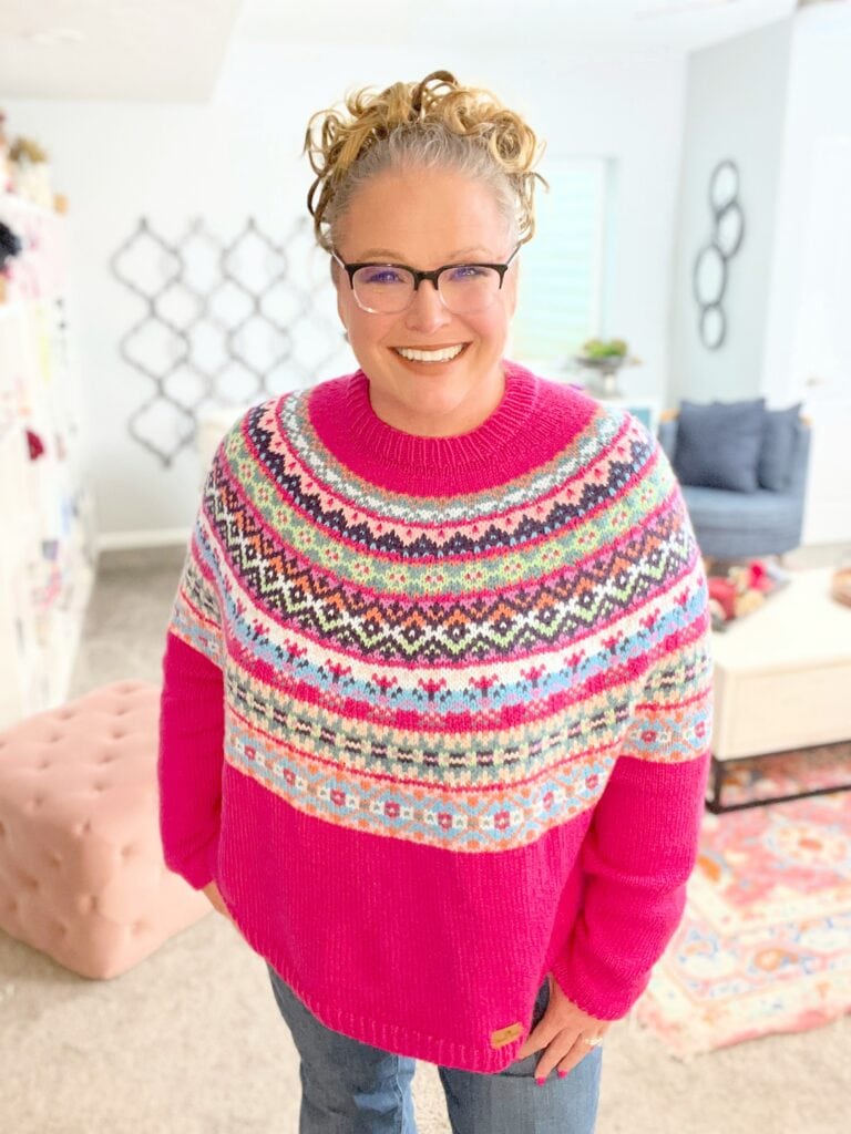 A woman with curly blonde hair, glasses, and a bright smile wears a colorful, patterned knit sweater—the knit and crochet versions of the same pattern are displayed in a cozy, well-lit room with light walls, a pink ottoman, and decorative accents. -Marly Bird