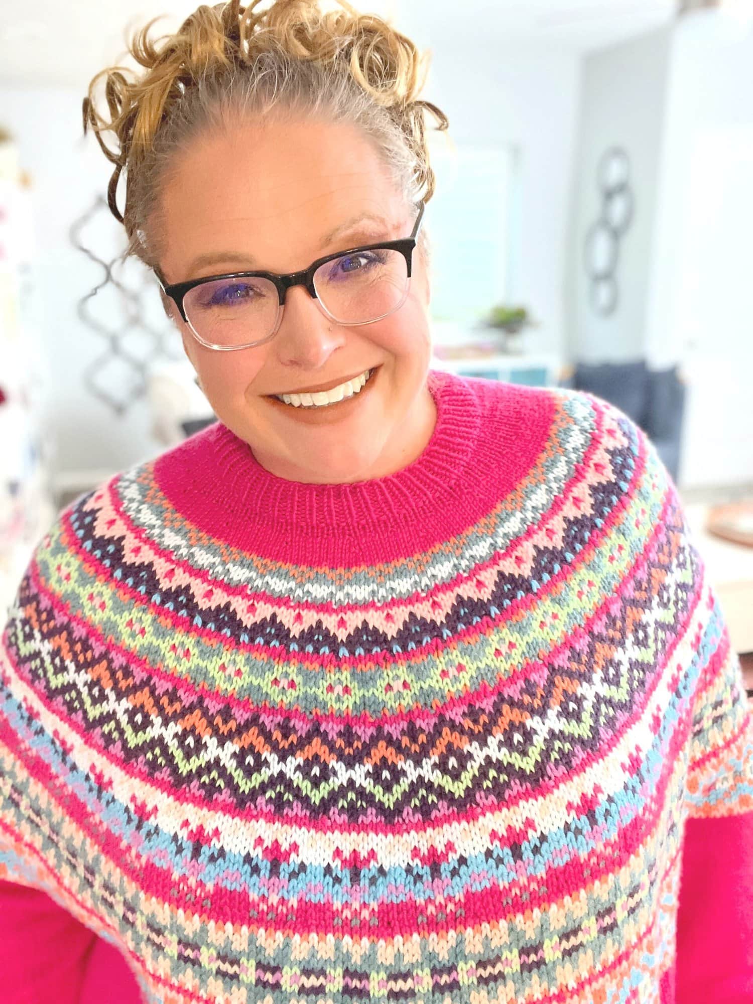 A smiling marly bird with curly hair, wearing glasses and a colorful, patterned knit sweater inspired by Free Colorwork stands indoors in a well-lit room. Prism Path Knit Swoncho -Marly Bird