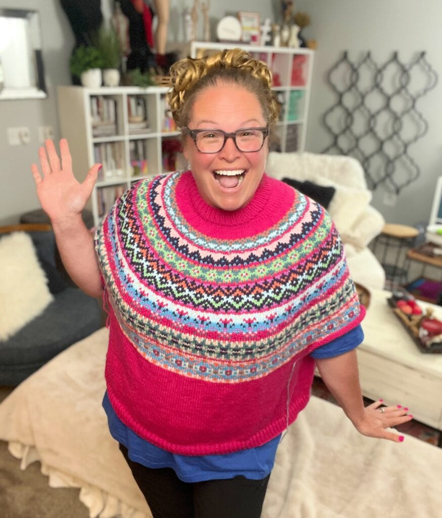 A smiling woman with curly hair and glasses waves with one hand. She is wearing a colorful, patterned knit poncho and standing in a cozy, well-lit living room with shelves and comfy furniture in the background. -Marly Bird