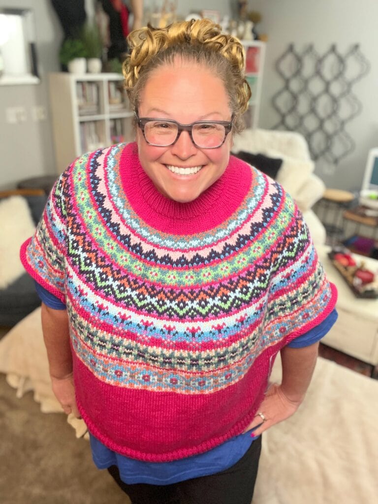 A smiling person with glasses and curly hair wears a brightly colored Prism Path Knit Swoncho. They stand indoors in a cozy room with bookshelves, a chair, and decor visible in the background. -Marly Bird