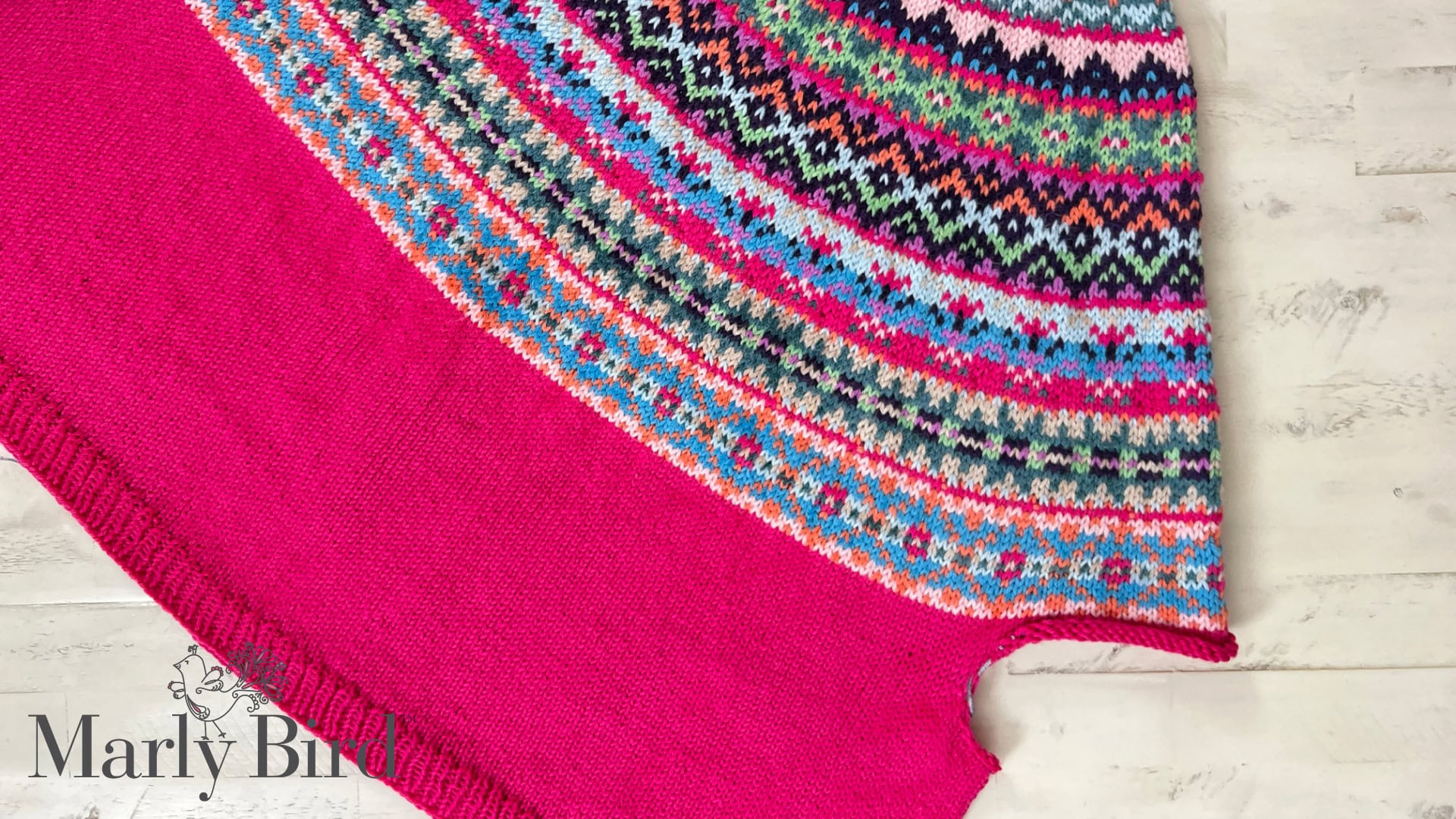 A close-up of a colorful knitted sweater with intricate patterns and vibrant shades of pink, blue, green, black, and white. The lower part is solid bright pink. "Marly Bird" is written at the bottom left. -Marly Bird