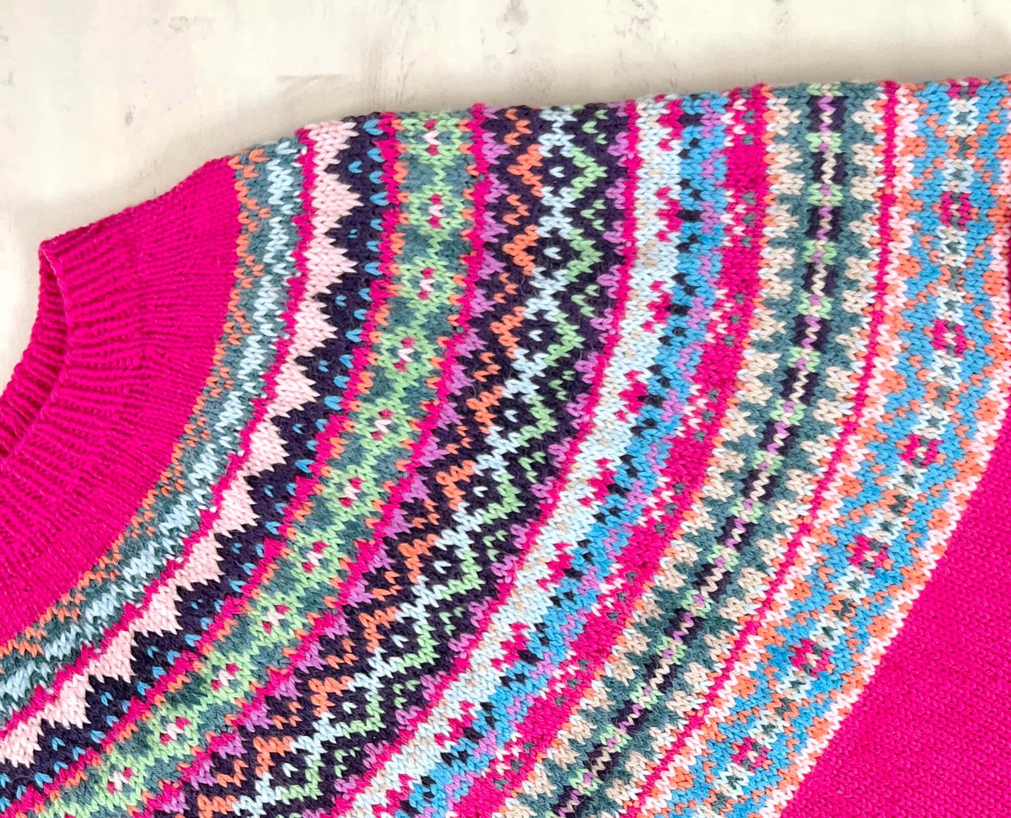 A close-up of the Prism Path Knit Swoncho | Part 6 showcases a pink-based sweater adorned with vibrant Fair Isle patterns in blue, green, orange, black, and white around the yoke. -Marly Bird