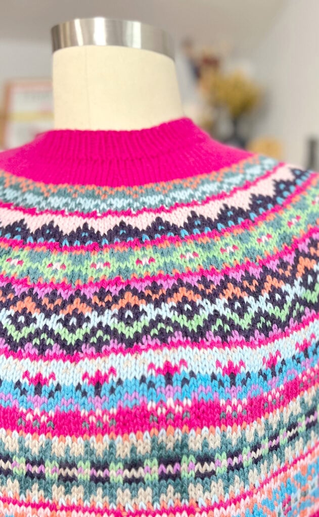 A close-up of the Prism Path Knit Swoncho Part 6 showcases a colorful knit sweater with intricate patterns in pink, blue, green, black, and white on a mannequin, featuring a bright pink ribbed collar. -Marly Bird