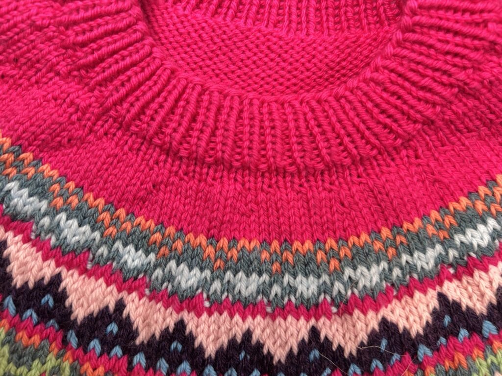 A close-up of a knitted sweater from the Prism Path Knit Swoncho, with a ribbed, bright pink collar and a colorful pattern of zigzag and diamond shapes in green, black, pink, orange, and white. Free Pattern for the Prism Make-Along 2025!. -Marly Bird