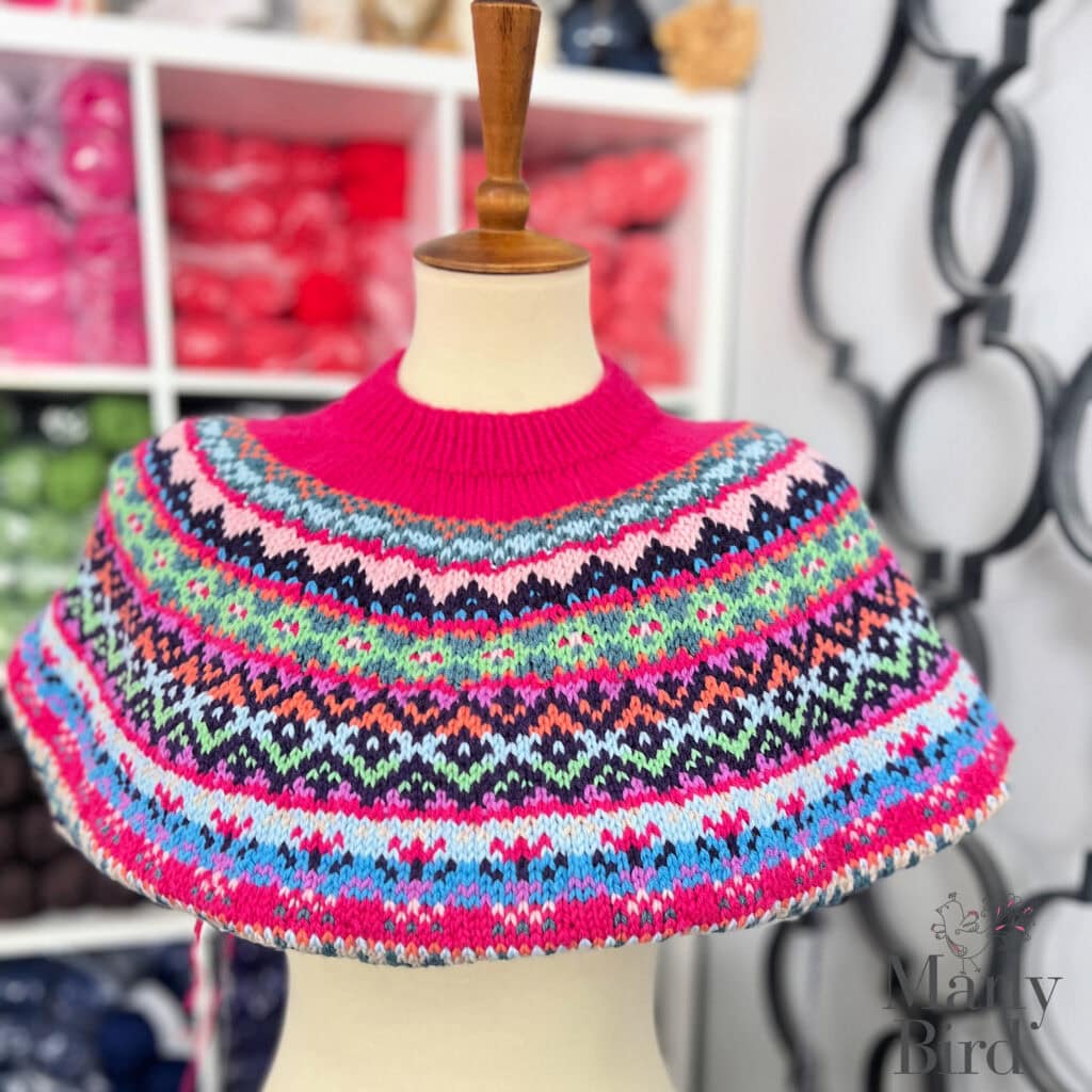 A mannequin displays a colorful, hand-knitted yoke with intricate, multicolored geometric patterns from the Prism Path Knit Swoncho. Yarn shelves in pink and other hues are visible in the blurred background. -Marly Bird