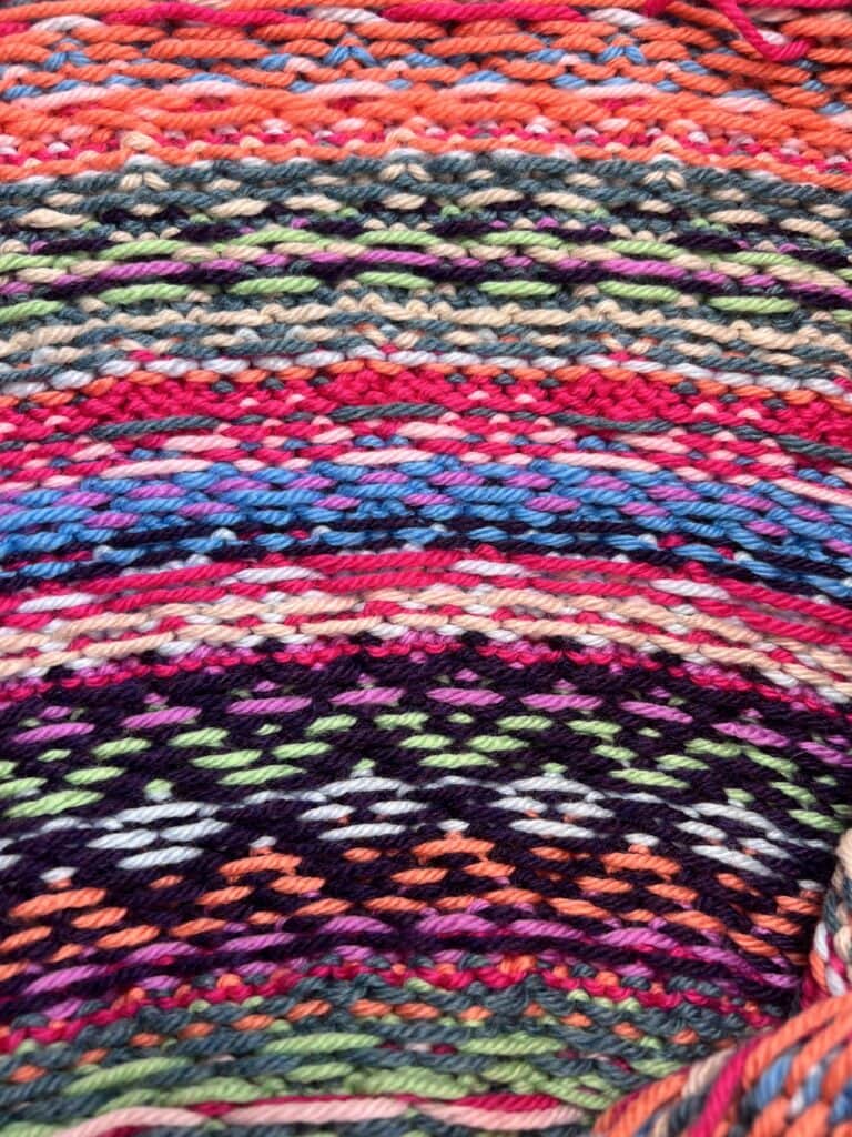 Close-up of a colorful knitted fabric, part of the Prism Path Knit Swoncho Free Pattern, featuring horizontal stripes and intricate geometric patterns. The vibrant texture and stitching are clearly visible—a perfect inspiration for your Make-Along 2025. -Marly Bird