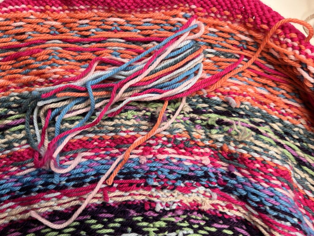 Close-up of a colorful knitted fabric from the Prism Path Knit Swoncho, with loose yarn ends in vibrant pink, orange, blue, green, and purple—showing the back side of this Free Pattern for the Prism Make-Along 2025. -Marly Bird