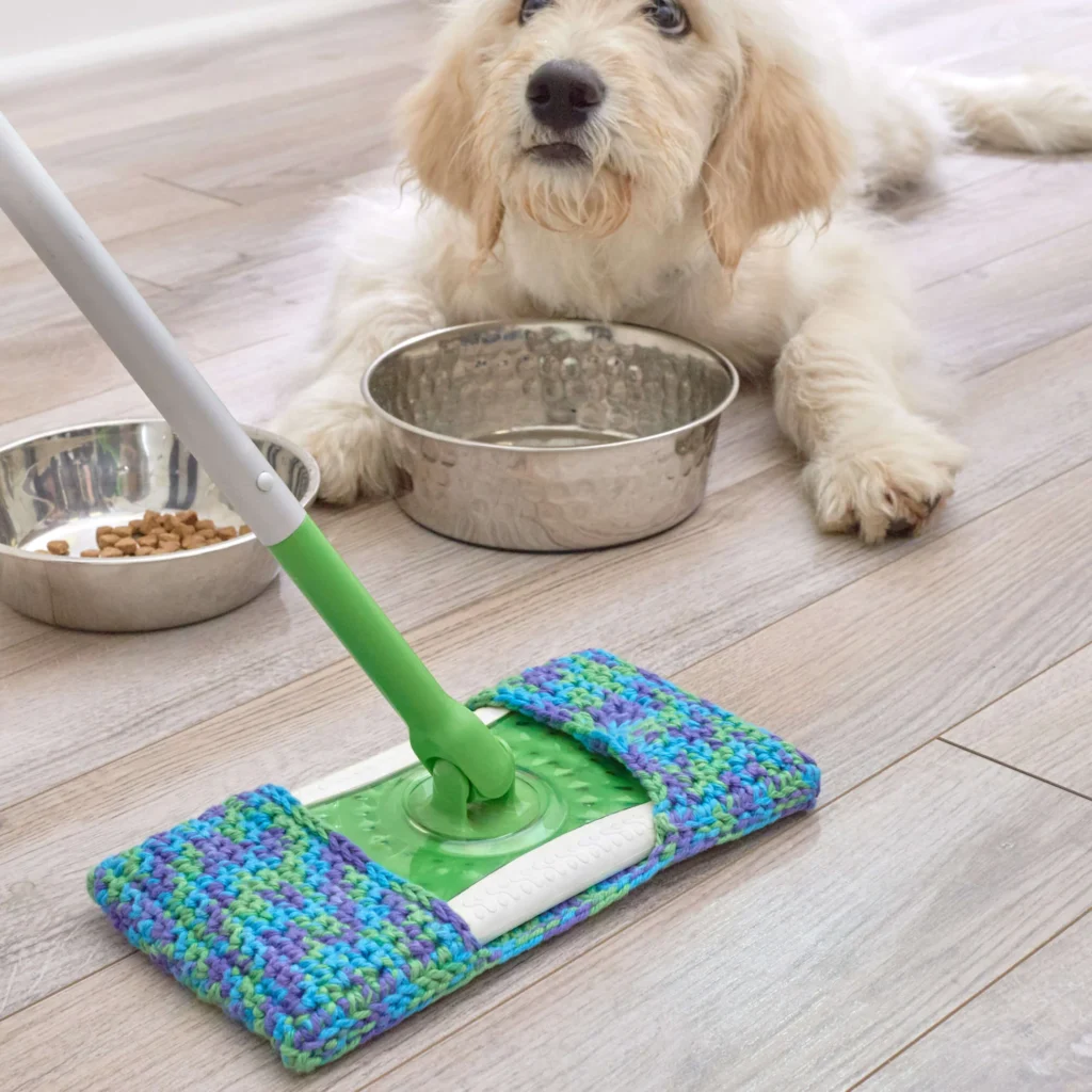 A fluffy dog relaxes on a wooden floor beside two bowls, one empty and one with food. In front of the dog, a mop with a colorful, crocheted cover awaits spring cleaning duties. The scene is well-lit and tidy. -Marly Bird