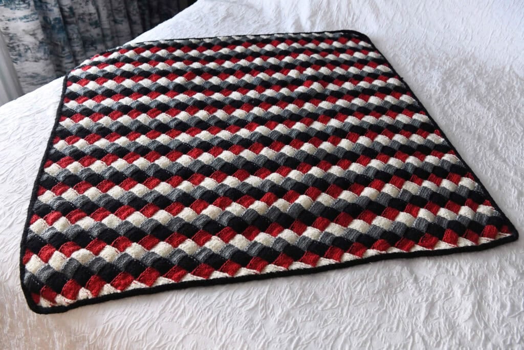 A handmade, rectangular quilt with a diagonal checkerboard pattern in black, red, and white rests on a white textured bedspread—perfect as inspiration for an entrelac knit baby blanket or a unique knit baby blanket gift. -Marly Bird