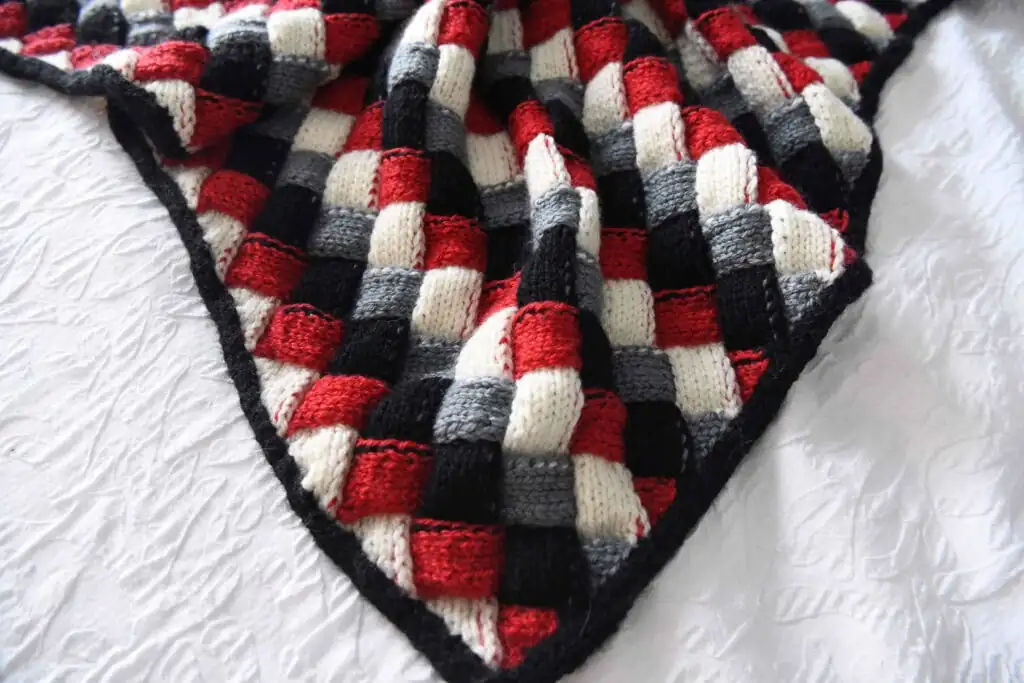 A knitted blanket with a checkerboard pattern in red, black, gray, and white squares is spread over a textured white surface. This cozy entrelac knit baby blanket appears soft and has a striking black border. -Marly Bird