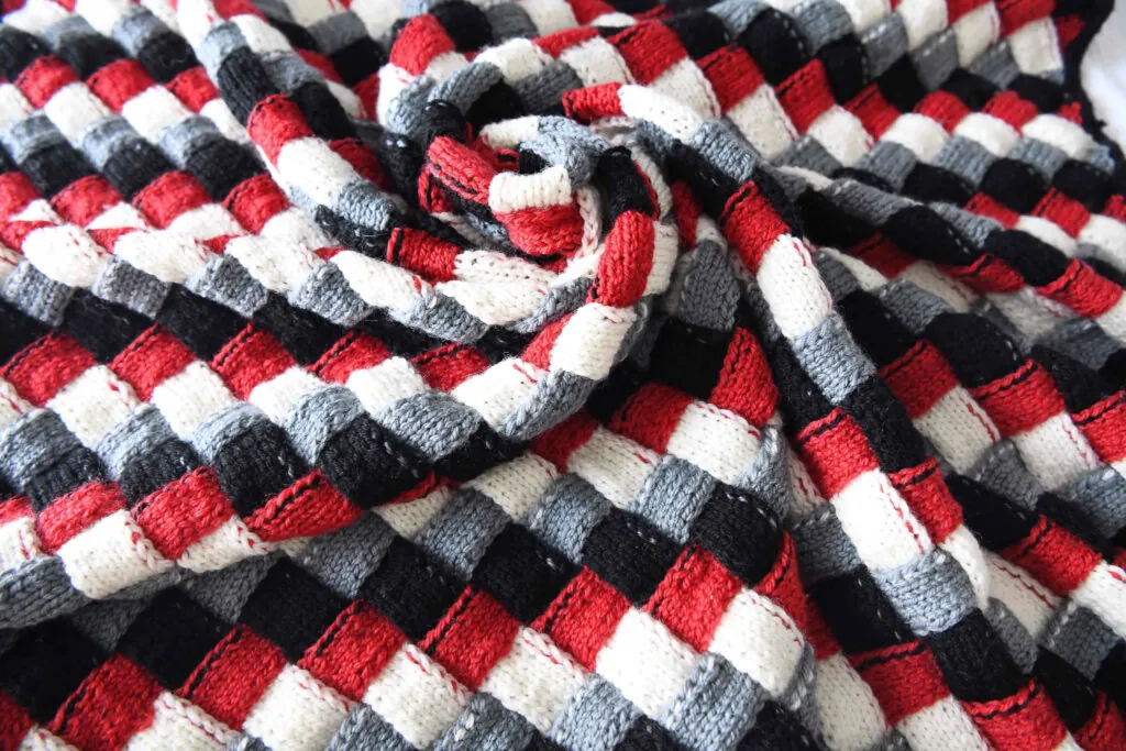 A knitted entrelac baby blanket with a checkered pattern in black, red, white, and gray, partially rolled and spread out, showing its textured stitches and vibrant colors. -Marly Bird