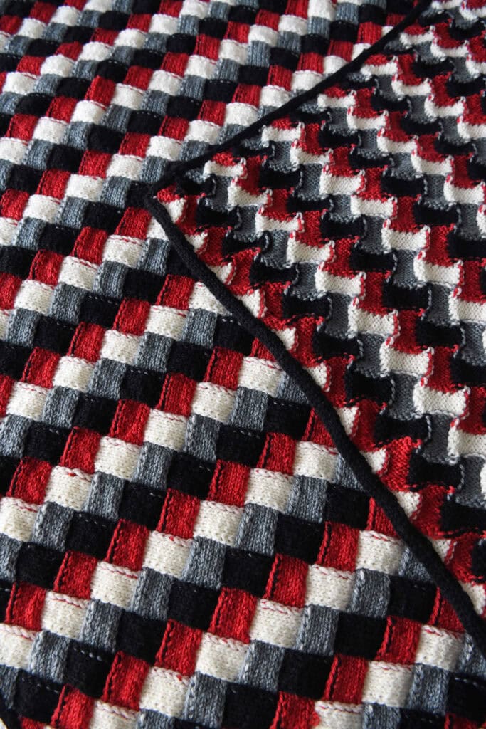 A close-up of a textured, woven fabric featuring a geometric checkerboard pattern in red, black, gray, and white squares—resembling an entrelac knit baby blanket—with one corner of the fabric folded over. -Marly Bird