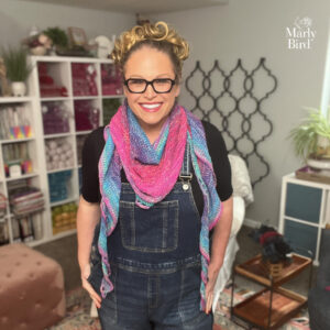 Person wears a colorful pink and blue knit scarf with visible chunky stitch texture, standing in a yarn-filled craft room.