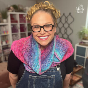 Woman models a vibrant crochet crescent shawl in pink, blue, and purple, showing off stitch texture and drape.