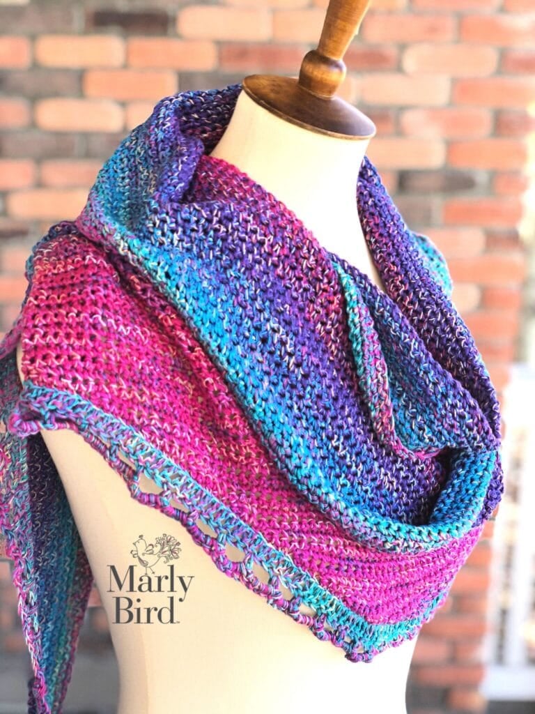 Hand-knit crescent shawl in purple, blue, and pink with visible stitch detail draped on a mannequin; blurred brick wall behind.