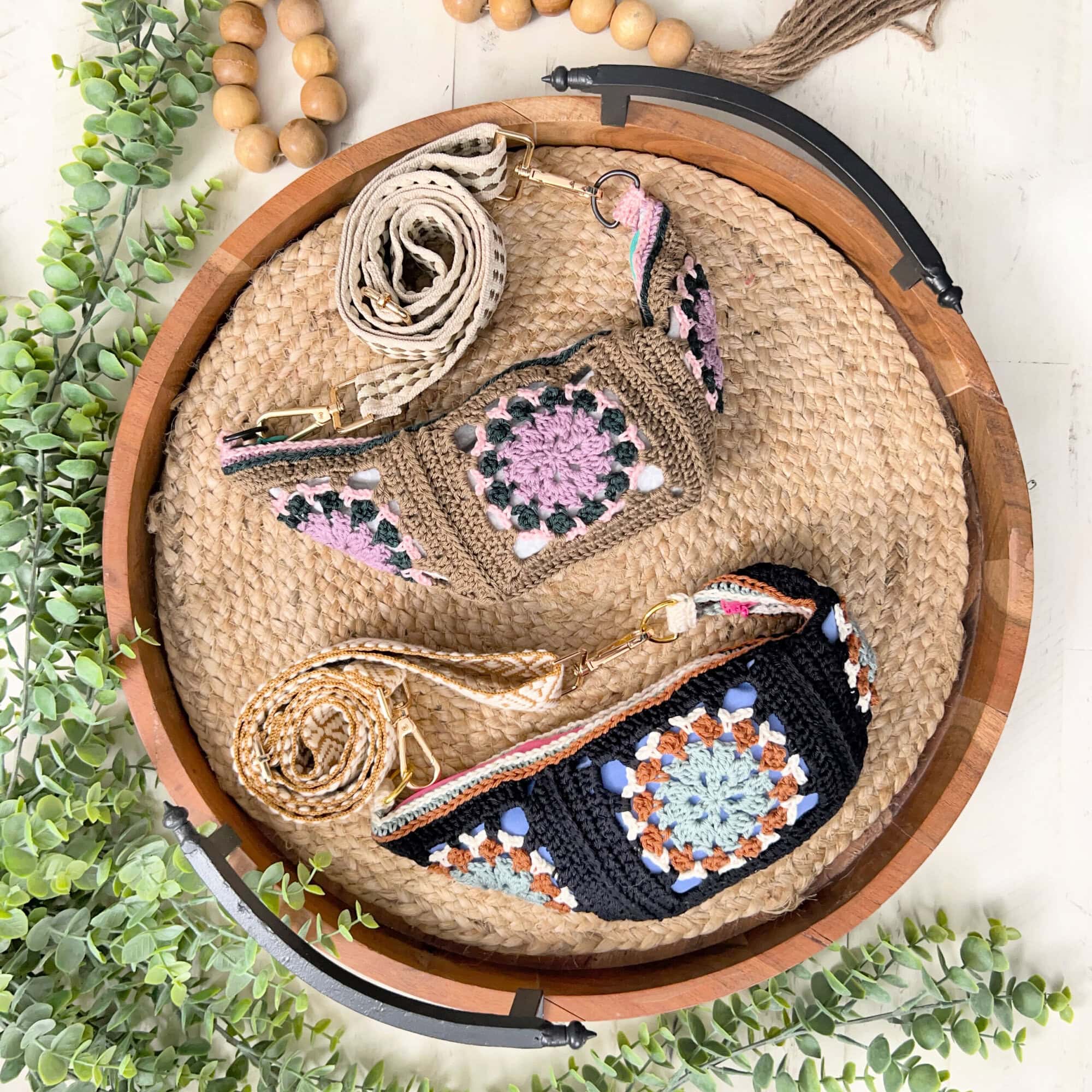 Two crocheted bags with colorful granny square patterns and woven straps, including a Crochet Crossbody Bag Pattern, are arranged on a round woven tray, surrounded by greenery and wooden bead garlands on a light surface. -Marly Bird